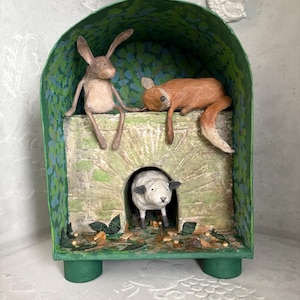 May include: A charming diorama depicting a brown rabbit and a red fox relaxing on a stone structure, with a sheep in an arched doorway. The structure is painted green with leaf patterns, and a white bird sculpture is mounted above.