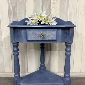 May include: A blue corner table with a single drawer and a lower shelf. The drawer has a bird-shaped pull and a painted floral design. The table is decorated with white flowers.