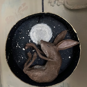 May include: A round, dark blue ornament with a gold rim, featuring a brown hare reaching for a white moon against a starry sky. The ornament is suspended by a dark blue string, creating a whimsical, celestial theme.