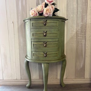May include: A sage green, oval cabinet with four drawers and bunny-shaped drawer pulls. The cabinet has curved legs and is topped with pink roses. The cabinet is approximately 76 cm tall.