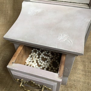 May include: A small, antique-style jewelry box in a soft lavender hue. The box features a drawer with a brass handle and is filled with a strand of white pearls. The top has a mirror and floral carvings.