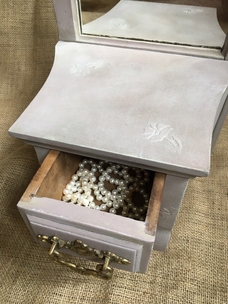 May include: A small, antique-style jewelry box in a soft lavender hue. The box features a drawer with a brass handle and is filled with a strand of white pearls. The top has a mirror and floral carvings.