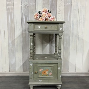 May include: A sage green bedside table with a drawer and cabinet, decorated with floral and paw print accents. A painted fox scene decorates the cabinet door. A bouquet of pink roses rests on top. The table is approximately 76 cm tall.