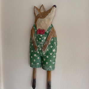 May include: Wooden fox figurine with brown fur, wearing green overalls with white flower accents and a red bow tie. The fox has a whimsical, hand-carved appearance, with its head tilted upwards. The figurine is mounted on a white wall.