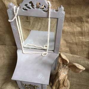 May include: A gray vanity mirror with a small drawer and a pearl necklace draped over it. A ceramic rabbit figurine sits to the right. The vanity has floral carvings and a brass drawer pull. The background is a burlap fabric.