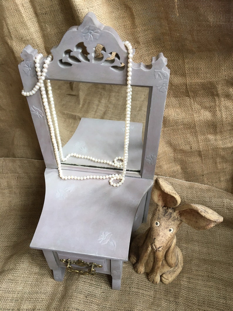 May include: A gray vanity mirror with a small drawer and a pearl necklace draped over it. A ceramic rabbit figurine sits to the right. The vanity has floral carvings and a brass drawer pull. The background is a burlap fabric.
