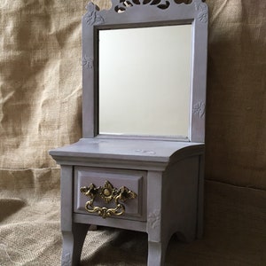 May include: A small, gray vanity with a mirror, drawer, and decorative gold-colored handle. The mirror is framed with ornate carvings, and the vanity has a classic, vintage design. The vanity is painted in a muted gray color.
