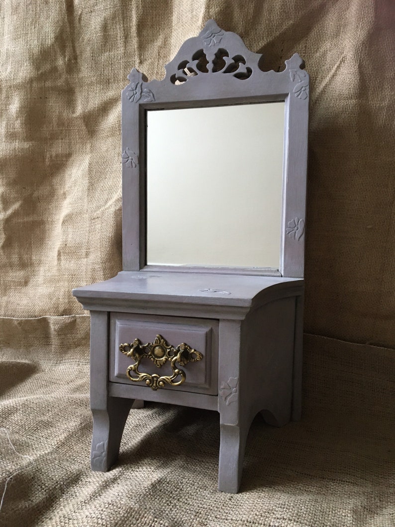 May include: A small, gray vanity with a mirror, drawer, and decorative gold-colored handle. The mirror is framed with ornate carvings, and the vanity has a classic, vintage design. The vanity is painted in a muted gray color.
