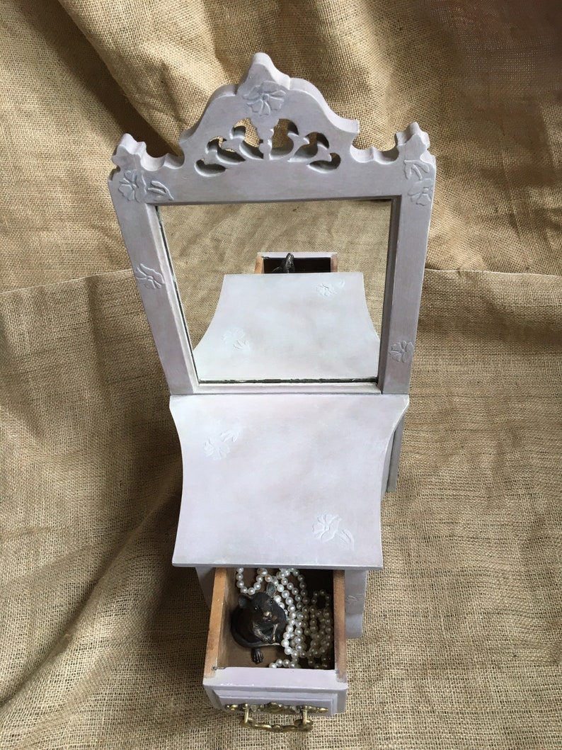 May include: A light purple jewelry box with a mirror and a drawer. The box has a decorative floral design on the top and sides. The drawer is open, revealing a pearl necklace and other jewelry. The box is made of wood and has a brass handle.