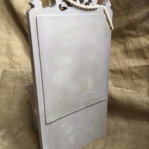 May include: A light gray, wooden jewelry display with a decorative top and a hinged front panel. A strand of white pearls is draped across the top. The display is set against a textured, tan background.