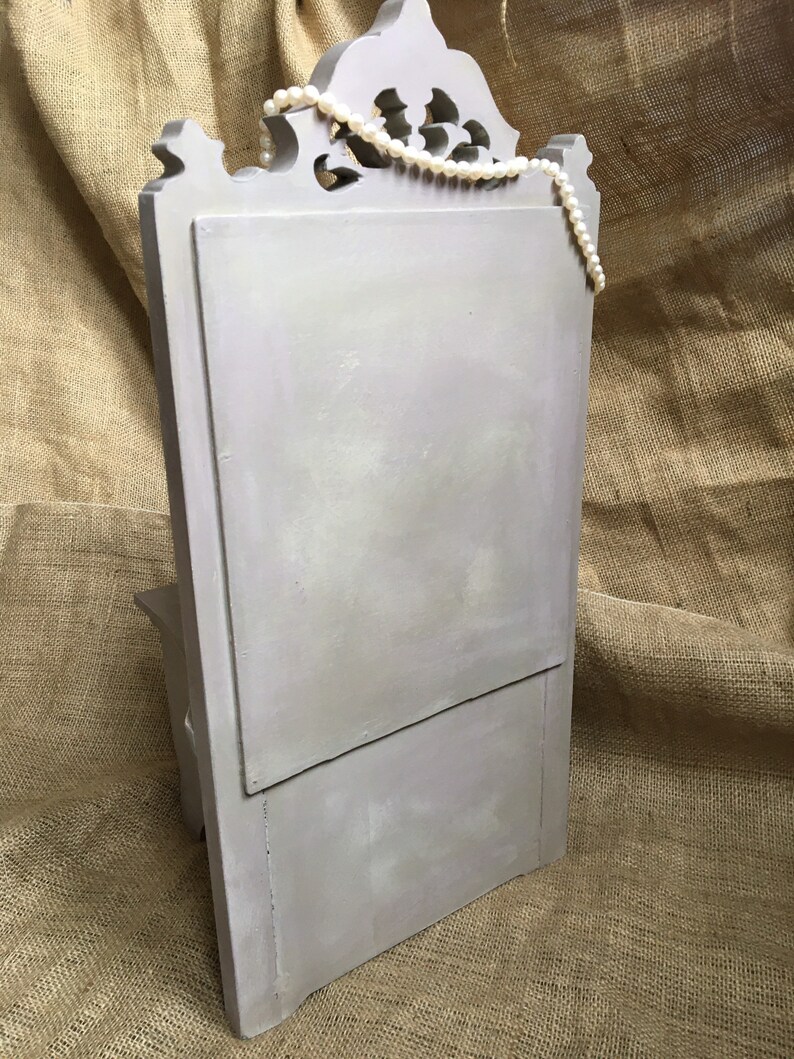 May include: A light gray, wooden jewelry display with a decorative top and a hinged front panel. A strand of white pearls is draped across the top. The display is set against a textured, tan background.