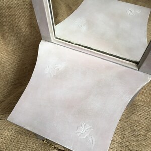 May include: A vintage-style vanity mirror with a light pink finish and floral embossed details. The mirror has a rectangular shape with a curved base and a small brass handle. The mirror is designed to sit on a table or dresser.