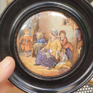 May include: A small, round framed print with a black frame. The print shows a group of children and a woman in a room. The woman is seated and appears to be knitting. The children are interacting with her. The print has a vintage aesthetic.