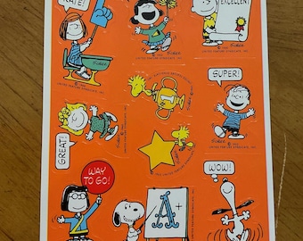 Könnte beinhalten: Ein Satz von neun Vintage-Peanuts-Charakteraufklebern auf orangefarbenem Hintergrund. Die Aufkleber zeigen verschiedene Peanuts-Charaktere, darunter Snoopy, Charlie Brown, Lucy und Sally. Die Aufkleber haben verschiedene Sätze darauf, wie z. B. "First-Rate!" und "Hurray!"
