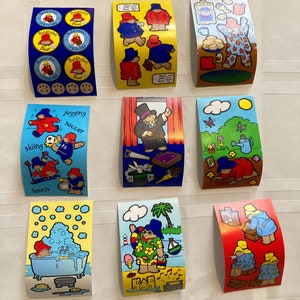 Puede incluir: Una colección de pegatinas coloridas de Paddington Bear. Las pegatinas muestran a Paddington en varias escenas, incluyendo practicando deportes, tomando un baño y disfrutando de unas vacaciones. Algunas pegatinas tienen texto como "Paddington Bear" y "P&Co 1985".