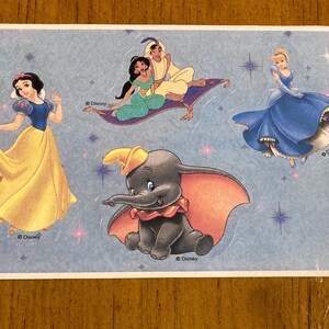 May include: A sheet of Disney character stickers featuring Snow White in a yellow dress, Jasmine and Aladdin on a magic carpet, Cinderella in a blue gown, and Dumbo the elephant. The background is light blue with star accents.