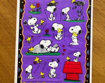 Può includere: Un foglio di adesivi rettangolare con diverse illustrazioni di Snoopy e Woodstock su sfondo viola. I personaggi sono raffigurati in varie pose, tra cui seduti su una cuccia rossa, mentre dormono e interagiscono con fiori e nidi.
