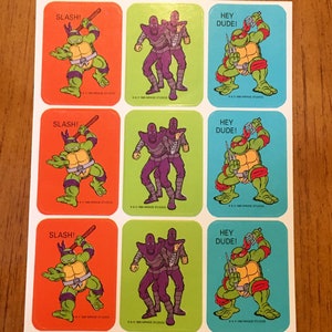 May include: Nine vintage Teenage Mutant Ninja Turtles stickers. Three stickers feature a green turtle with a red bandana, wielding a katana and saying "Slash!" Three stickers feature two purple figures with masks and saying "Hey Dude!" Three stickers feature a green turtle with a blue bandana, wielding a katana and saying "Hey Dude!"