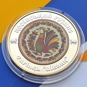 Puede incluir: Una moneda dorada dentro de un soporte de plástico transparente. La moneda presenta una colorida ilustración de un gallo rodeado de motivos florales y texto en un diseño circular. El borde de la moneda tiene texto adicional.
