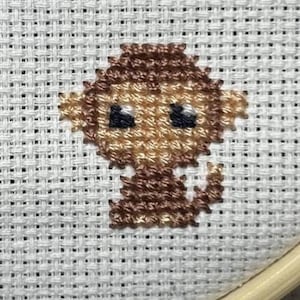 Può includere: Ricamo a punto croce di una piccola scimmia nei toni del marrone e beige, con occhi neri, su tessuto bianco. Il ricamo è posto in un telaio di legno, che mostra un progetto artigianale.