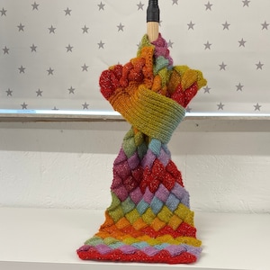 Puede incluir: Una bufanda de punto colorida con un patrón de arcoíris se muestra en un  de madera. La bufanda está hecha con un hilo grueso y tiene un tejido texturizado. La bufanda se muestra en un estante con varios ovillos de lana de varios colores.