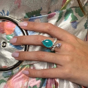 Puede incluir: Primer plano de un anillo con una gran piedra preciosa turquesa en forma de pera. El anillo tiene una banda plateada con piedras blancas más pequeñas y una piedra rosa más pequeña en forma de pera. El anillo está en una mano.
