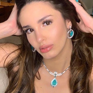 Puede incluir: Una mujer con un conjunto de joyas de turquesa y diamantes. El conjunto incluye un collar con un colgante en forma de lágrima, pendientes a juego y un anillo. Las joyas destacan sobre el cabello castaño de la mujer.