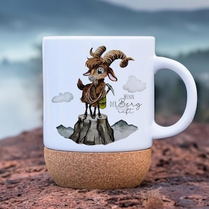 Può includere: Tazza in ceramica bianca con base in sughero. Presenta un cartone animato di una capra su una montagna, con la scritta "Wenn der Berg ruft". La capra è marrone con grandi corna e una piccola borsa verde. Ideale per bevande calde.