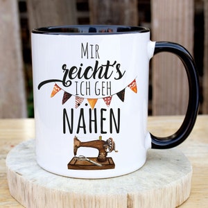 Puede incluir: Taza de cerámica blanca con borde y asa negros. La taza tiene un gráfico negro que dice "Mir reicht's ich geh nähen" con una máquina de coser vintage y una hilera de banderines coloridos.