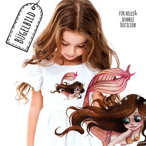 Könnte beinhalten: Ein weißes T-Shirt mit zwei Bügelbildern von Meerjungfrauen mit langen braunen Haaren und rosa Schwänzen. Der Text "Für helle & dunkle Textilien" ist rechts im Bild gedruckt.
