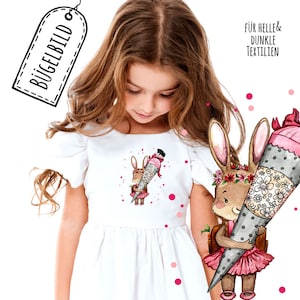 Può includere: Abito bianco con un'illustrazione di un coniglietto che tiene un cono scolastico. Il coniglietto indossa un vestito rosa e una corona di fiori. L'immagine include il testo "BÜGELBILD" su un'etichetta.