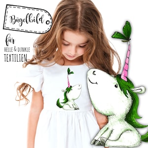 May include: A white dress with a green and pink unicorn design. The unicorn has a green mane and tail, a pink horn, and a green leaf on its head. The text "Bügelbild für helle & dunkle Textilien" is on a white label.