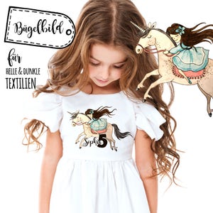 Puede incluir: Una transferencia de planchado blanca con una niña montando un unicornio. El unicornio es marrón claro con una crin y una cola rosas. La niña lleva un vestido azul con una cinta rosa. La transferencia dice "Sophia 5".