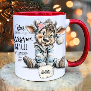 Op de afbeelding: Een witte keramische mok met een rode binnenkant en handvat. De mok heeft een cartoon hert in winterkleding en de tekst "Hier drin steckt Nordpol Magie Weihnachts-Tasse von Simone."