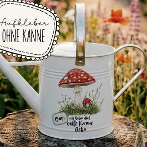 May include: White metal watering can with a silver handle and spout. The can features a red and white mushroom graphic and text in German. The watering can is sitting on a wooden stump in a garden setting.