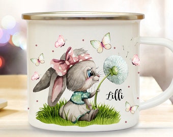 May include: White enamel mug with a watercolor illustration of a bunny holding a dandelion. The bunny is wearing a pink and white polka dot bow and a blue and white dress. The mug has the name "Lilli" written on it.