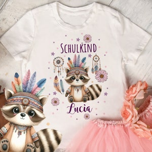 Könnte beinhalten: Weißes T-Shirt mit einem Cartoon-Waschbären, der einen bunten Kopfschmuck trägt und den Text "Schulkind Lucia".