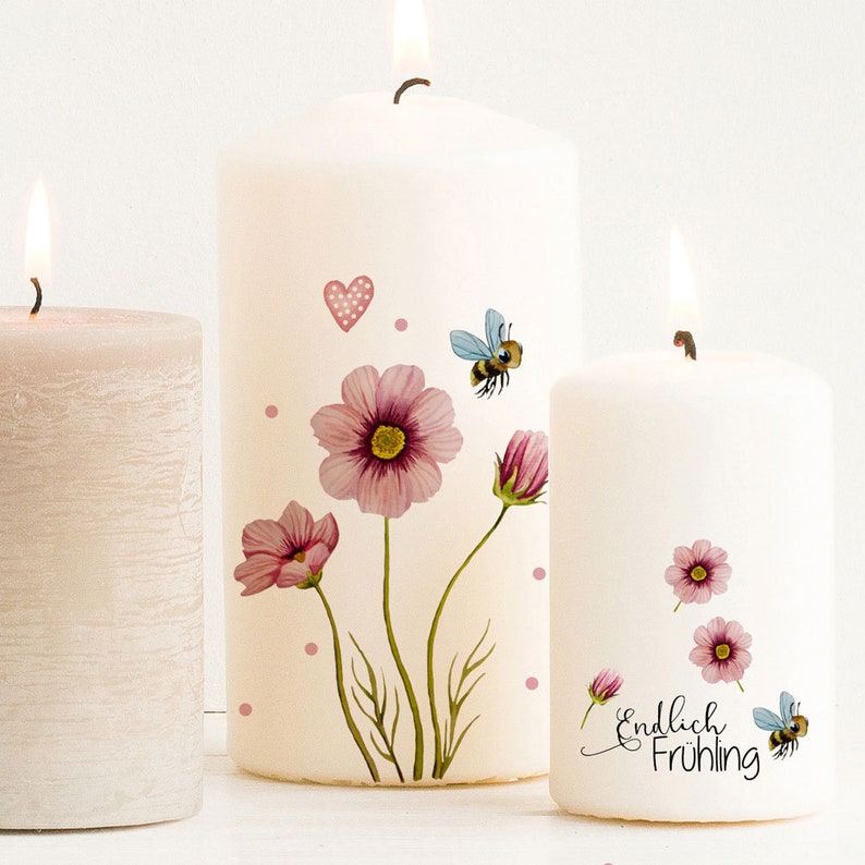 May include: Three lit pillar candles. The two front candles are white and decorated with pink flowers, bees, and hearts. The smallest candle has the words "Endlich Fr&uuml;hling" in black script. The third candle is a light pink color.