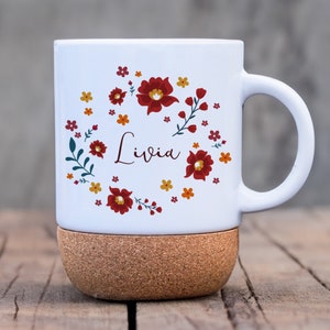 Könnte beinhalten: Weißer Keramikbecher mit einem floralen Design in Rot, Orange und Gelb, mit dem Namen "Livia" in Schreibschrift. Der Becher steht auf einem Korksockel, ideal für heiße oder kalte Getränke.