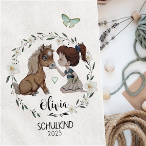 Puede incluir: Una ilustración de acuarela de una niña con cabello castaño y una camisa a rayas azul y blanca besando a un pony marrón. La ilustración está rodeada de una corona de flores blancas y hojas verdes. El texto "Olivia SCHULKIND 2023" está escrito debajo de la ilustración.