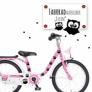 以下が含まれることがあります： 黒のフクロウのステッカーと黒のドットが付いたピンクの自転車。自転車は白いサドルと白いタイヤが付いています。看板には「Fahrradaufkleber "Eulen"」と書かれています