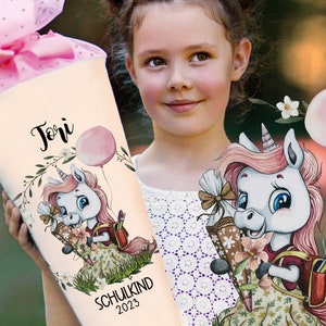 Puede incluir: Un cono escolar de unicornio blanco y rosa con el nombre "Jori" y el texto "Schulkind 2023". El cono está decorado con flores, hojas y un globo rosa.