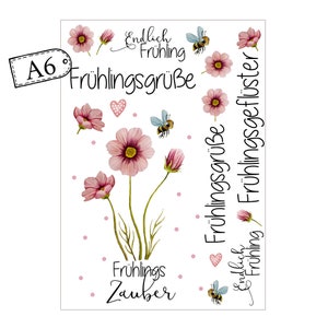 May include: A6-sized sticker sheet featuring watercolor illustrations of pink cosmos flowers, bees, and hearts. The sheet includes the German text "Fr&uuml;hlingsgr&uuml;&szlig;e" and "Fr&uuml;hlingszauber" in a decorative font, perfect for spring-themed crafts.