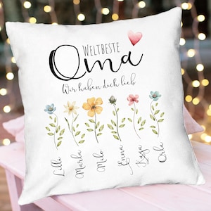May include: White pillow with the text "Weltbeste Oma" and "Wir haben dich lieb" in black lettering. A pink heart is above the text. Below the text are seven watercolor flowers with the names "Lilli", "Marlene", "Milo", "Emma", "Kaysa", "Leni", and "Orla" written below each flower.