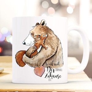 Może przedstawiać: Biały kubek ceramiczny z akwarelową ilustracją brązowego niedźwiedzia obejmującego wiewiórkę. Tekst na kubku brzmi "Weil mit Dir überall Zuhause ist."