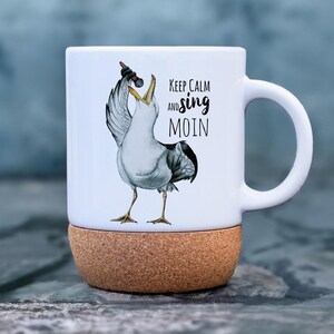 Op de afbeelding: Witte keramische mok met een kurken onderkant. De mok heeft een cartoon zeemeeuw die in een microfoon zingt met de tekst "Keep Calm and sing MOIN".