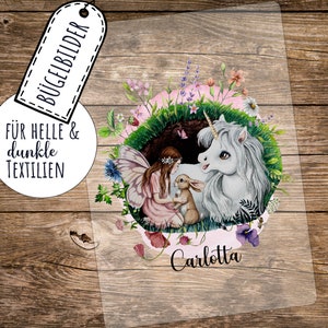 Puede incluir: Una lámina de transferencia transparente con una ilustración fantástica. Muestra un hada, un unicornio y un conejo en medio de flores y vegetación. El nombre "Carlotta" está escrito en cursiva. También se ve el texto "BÜGELBILDER" y "FÜR HELLE & dunkle TEXTILIEN".