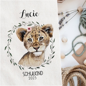 Op de afbeelding: Een aquarel-illustratie van een leeuwenwelpje met een roze bloemenkroon. Het welpje is omringd door een groene, bladerige krans. De tekst "Lucie" staat bovenaan de afbeelding en "Schulkind 2023" onderaan.