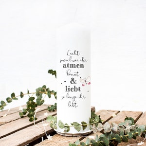 Könnte beinhalten: Weiße Kerze mit dem Text "Lacht soviel wie ihr atmen konnt & liebt so lange ihr lebt." Die Kerze ist mit einem kleinen Schmetterling und zwei rosa Beeren verziert.