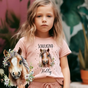 Könnte beinhalten: Ein pinkfarbenes T-Shirt mit einer Aquarell-Illustration eines Pferdes, das eine Blumenkrone trägt. Der Text "SCHULKIND" und "Holly" ist auf dem Shirt aufgedruckt.